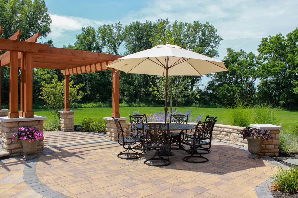 Open paver dining patio with umbrella table, pergola, stone wall, planter pots, ornamental grasses, and lawn view.
